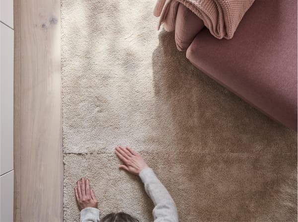 STOENSE low pile rug in off-white, seen from above in a living room, with a person feeling the rug’s soft, dense surface.