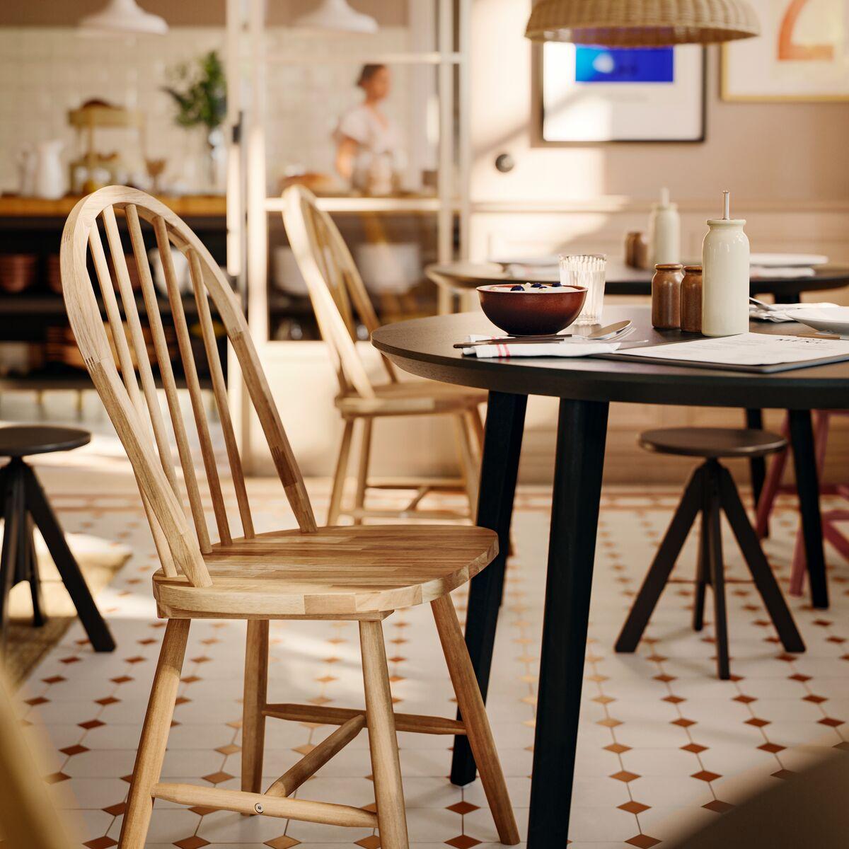 Stol SKOGSTA iz masivne akacije z obokanim naslonom za hrbet in vreteni stoji ob temni okrogli mizi v poslovnem jedilnem prostoru.