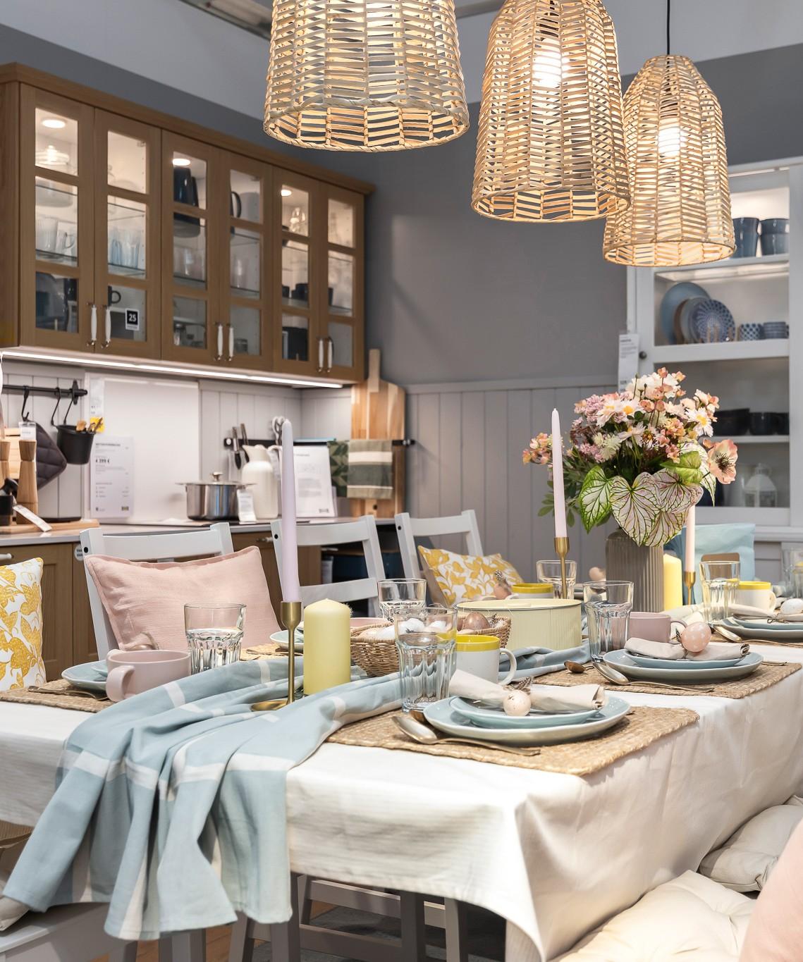 Столовая, сервированная к праздничному пасхальному ужину: стол с многослойным текстилем, посудой пастельных оттенков, стеклянной посудой, свечами и вазой с цветами. Над столом висят плетёные подвесные светильники, на заднем плане — шкафы и полки с предметами для дома.