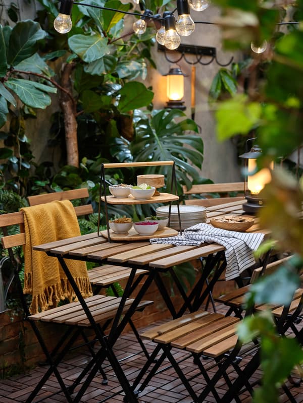 Stoly a stoličky TÄRNÖ stoja v záhrade na podlahe RUNNEN a sú osvetlené stolovou LED lampou STORHAGA a svetelnou LED reťazou UTSUND.