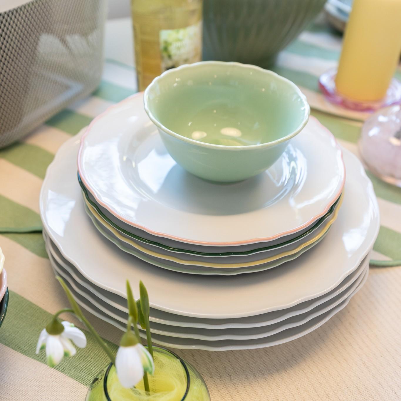 Стопка белых тарелок с пастельно-зелёной миской сверху, размещённая на зелёной полосатой подставке, рядом — весенние декоративные элементы.