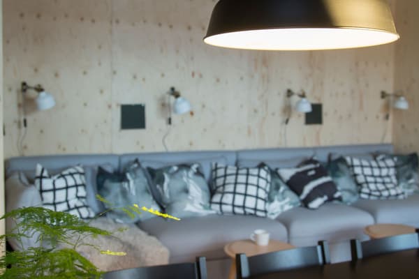 Stor grå soffa men svartvita kuddar i som står längs en ljus furuvägg. På väggen hänger vita lampor. 