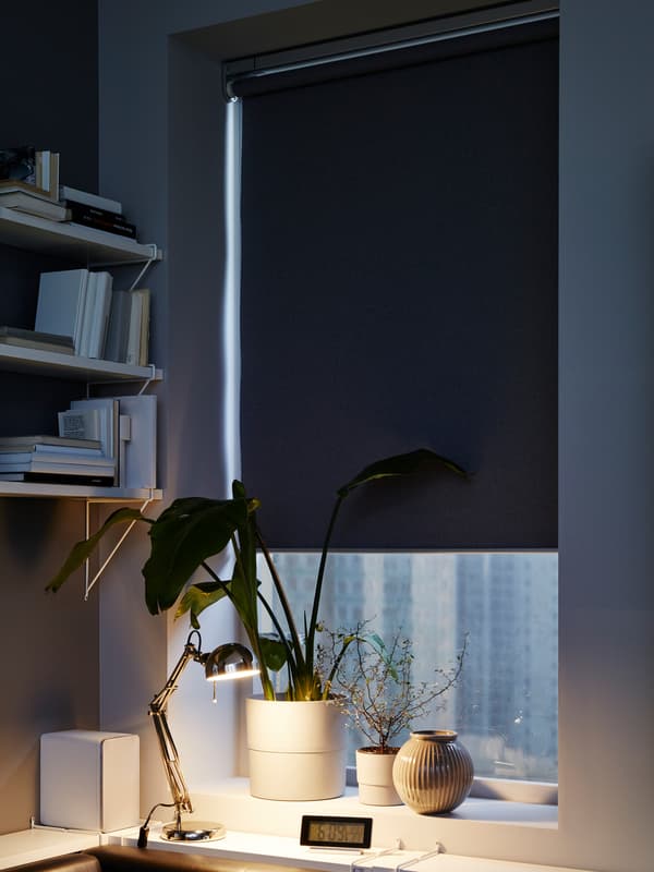 Store occultant FYRTUR couvrant la majeure partie d’une fenêtre, plantes en pots et vase sur l’appui de fenêtre et étagères murales blanches.
