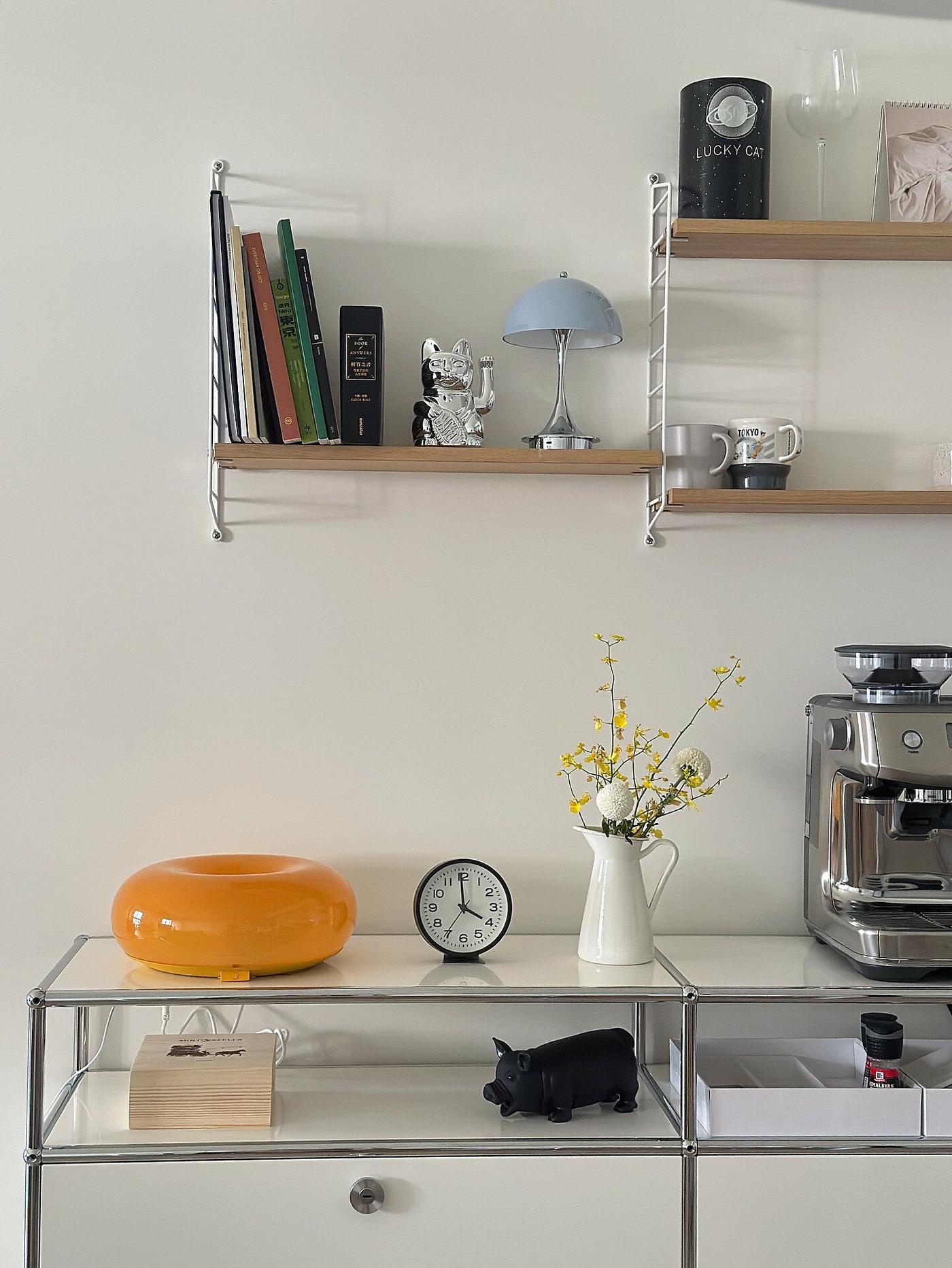 Strakke koffiecorner met een chromen espressomachine op een metalen dressoir, een gele decoratieve schaal, klok en bloemenvaas, wandplanken met boeken, mokken en accessoires, en een lichte wand als achtergrond.