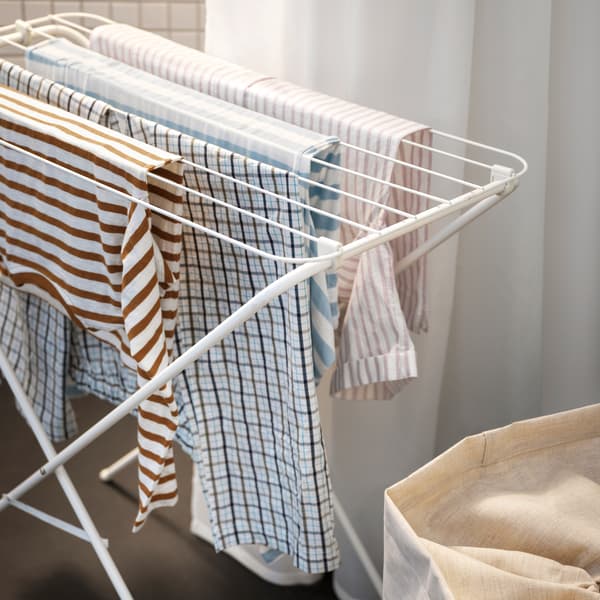 Striped and checked clothes hang on a white JÄLL drying rack in front of a white shower curtain, behind a beige laundry bag.