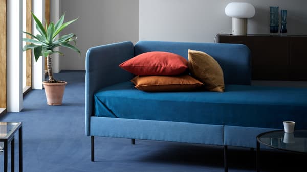 Struttura letto imbottita BLÅKULLEN abbinata a cuscini confortevoli in un soggiorno moderno ben illuminato. Accanto, una pianta con vaso e dietro un mobile con la lampada da tavolo DEJSA.