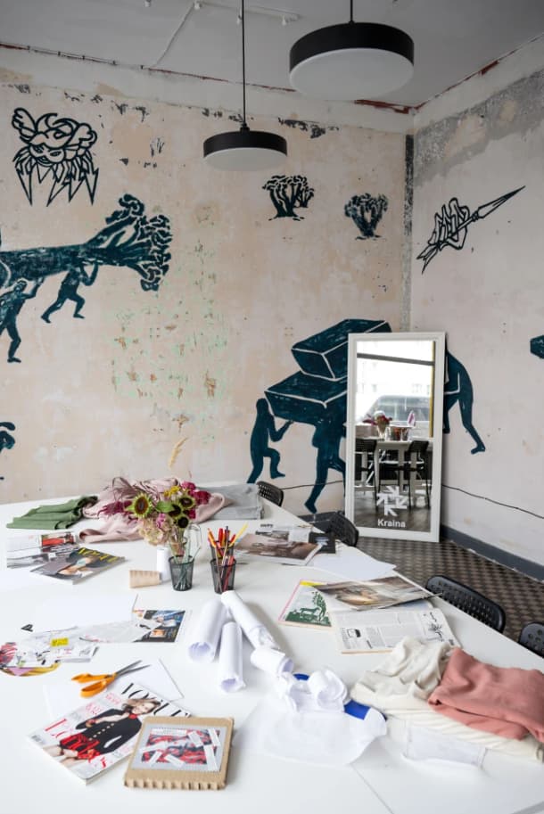 Studio projektowe Domu MoDy Kraina. W rodu widczone lustro stojace na ziemi  i oparte o ściane. Na głównym planie duży biały stół z tkaninami i szkicami