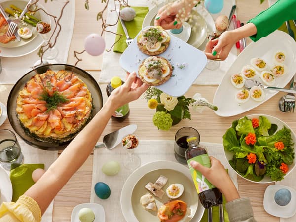 Stůl plný lahůdek na velikonoční hostinu, talíř s plněnými vajíčky, zapečené brambory s lososem, ruce si nad stolem podávají talířek s knäckebrotem s vajíčky a naloženými sledi.