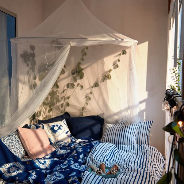 Sunlit balcony with double bed softly piled with cushions and blue-white bedlinen; mosquito net above, plants around.