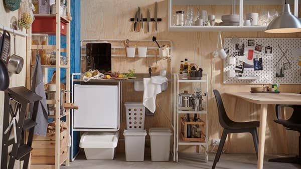SUNNERSTA mini-kitchen with accessories, an IVAR shelving unit and a table and chairs in a small room with plywood walls.