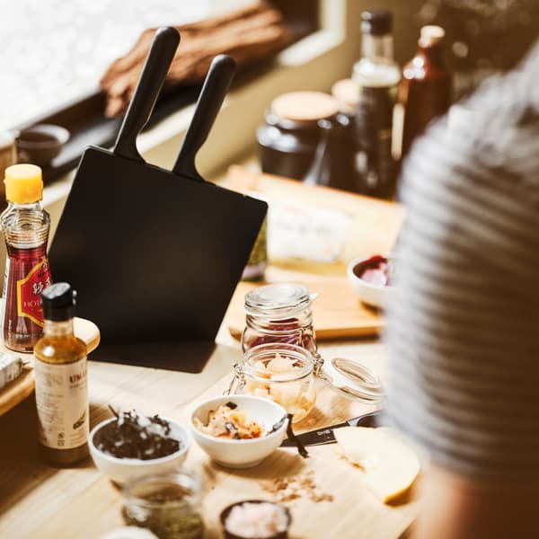 Superfície de mesa com suporte com facas JÄMFÖRA em preto, tábua de cortar APTITLIG, recipientes KORKEN e tigelas em branco.
