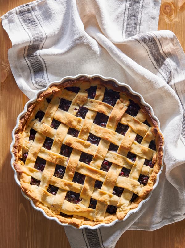 Suprafață din lemn cu o plăcintă cu afine cu împletitură de aluat la suprafață, proaspăt scoasă din cuptor, într-un platou de plăcintă VARDAGEN pe un șervet.