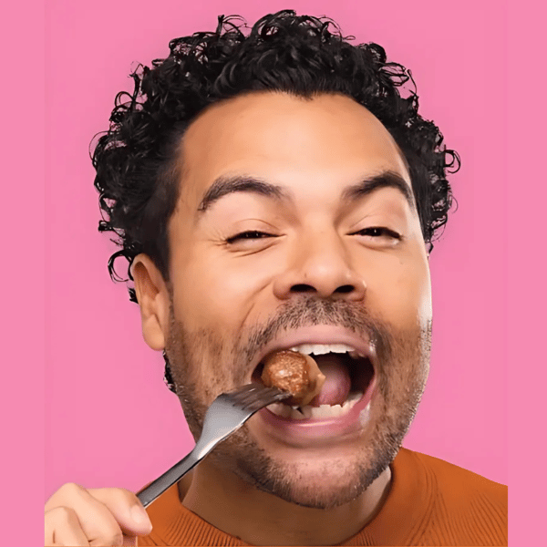 Sur cette image, on voit un homme souriant sur fond rose, prêt à croquer dans une boulette tenue avec une fourchette. L’ambiance est vive et gourmande, mettant en avant le plaisir de déguster les célèbres boulettes IKEA.