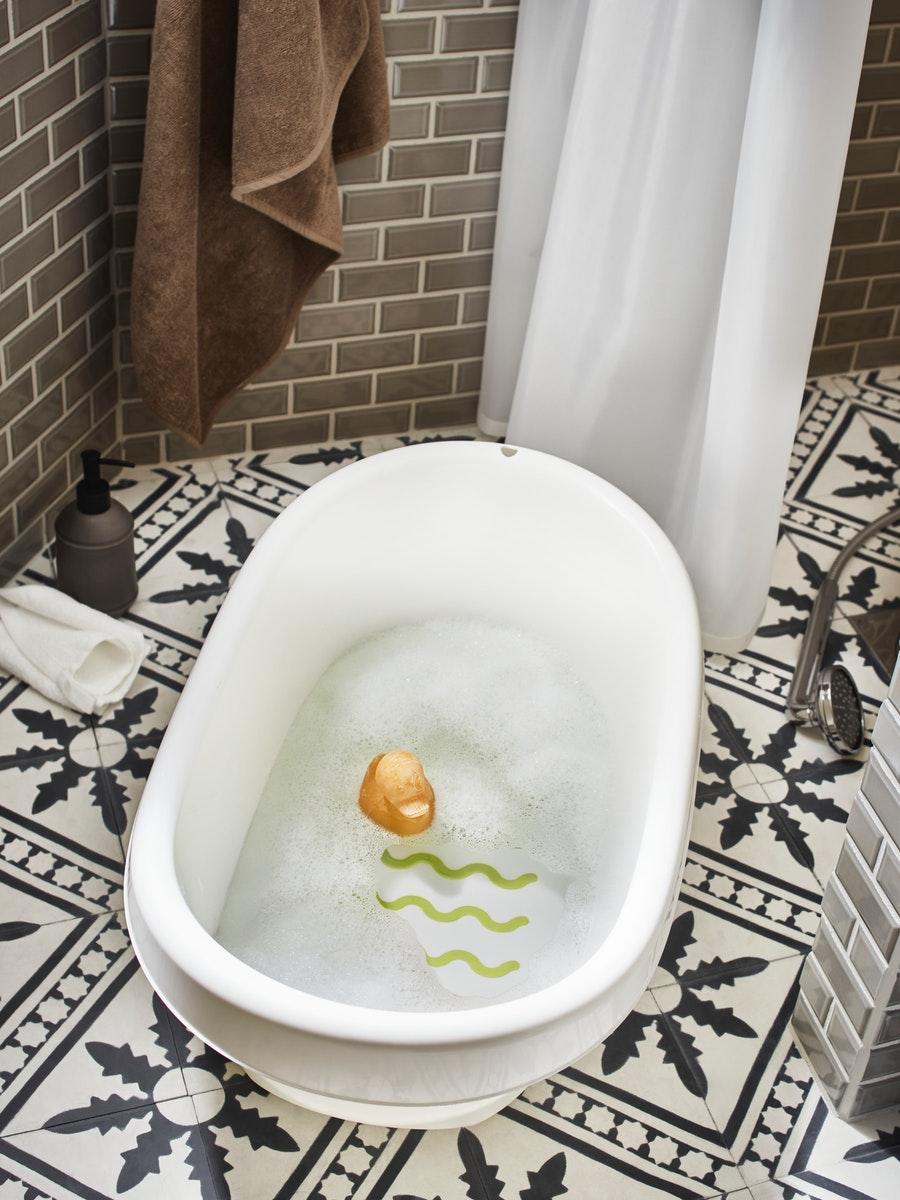 Sur le carrelage à motifs d’une salle de bain, une baignoire bébé LÄTTSAM en blanc/vert avec un canard en caoutchouc dans l’eau et une serviette de bain accrochée non loin.