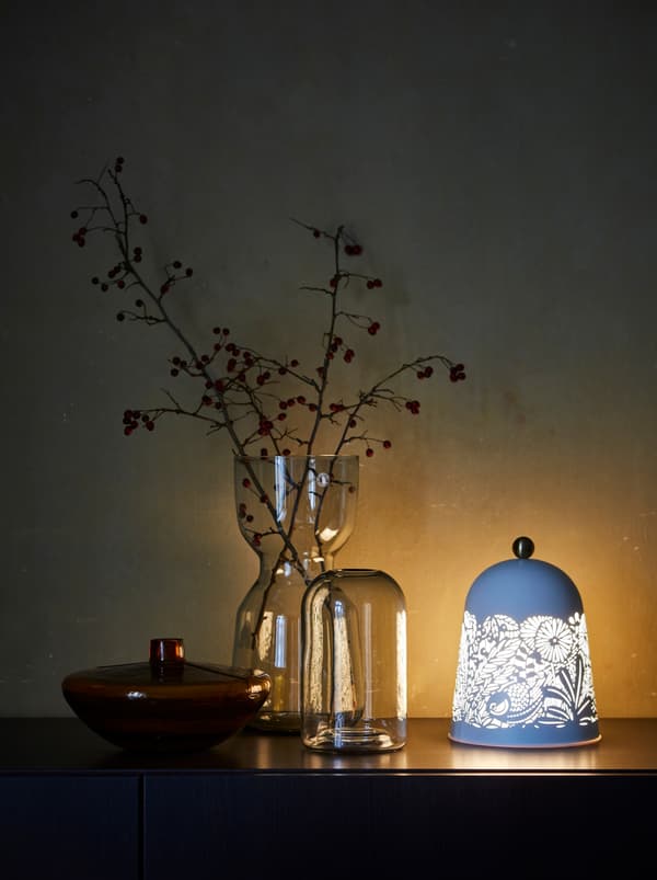 Sur un buffet, un grand vase décoratif orné de branchages d’automne, deux vases plus petits et une lampe aux motifs inspirés de la nature.