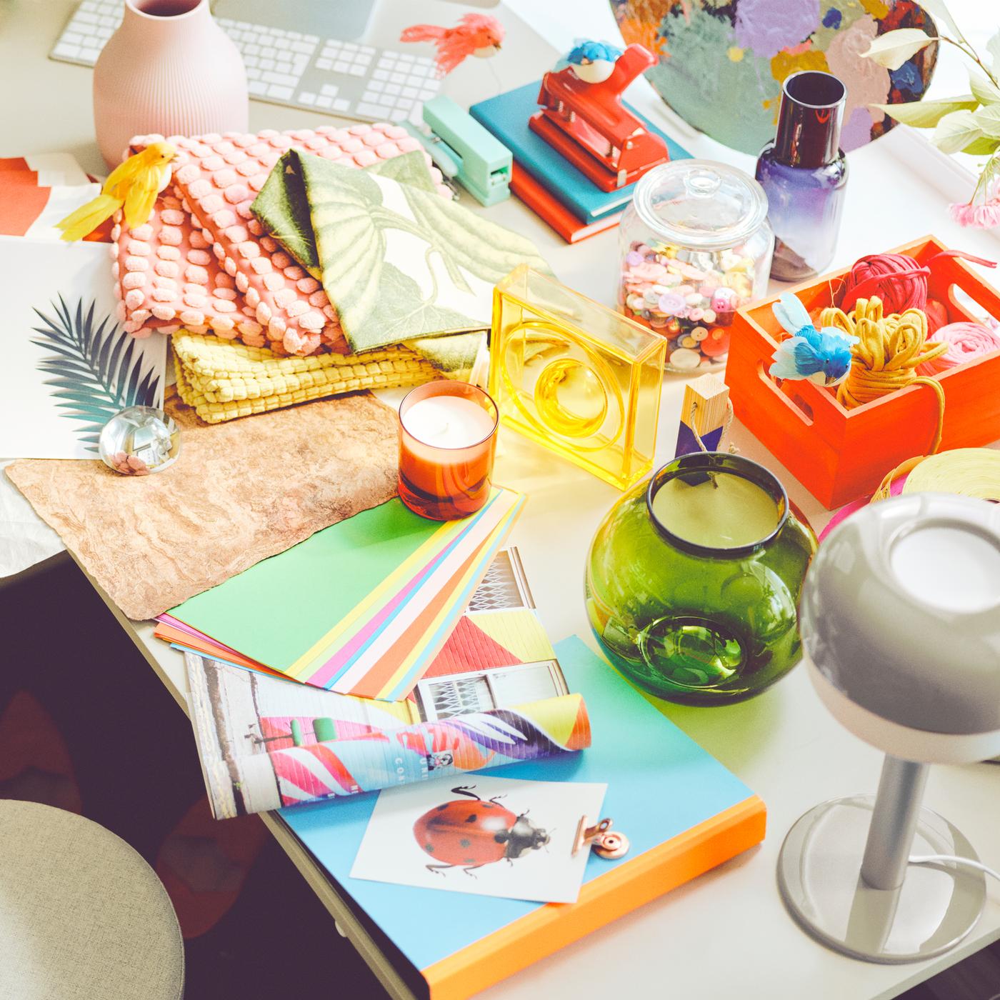 Sur un bureau sont posés des accessoires colorés épars, des vases, des housses de coussins et une lampe de table BLÅSVERK beige.