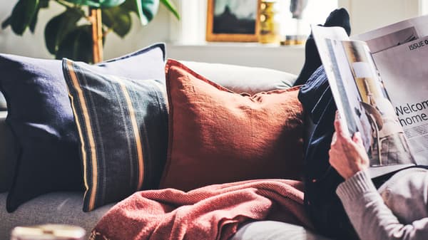 Sur un canapé, coussins dans des housses STORTIMJAN et DYTÅG à côté d’une personne lisant un magazine.