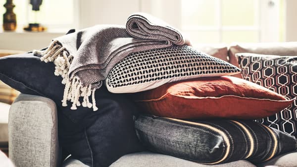 Sur un canapé gris, une pile de coussins dans des housses KUSTFLY et DYTÅG et un plaid.
