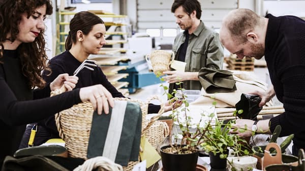 Sur un grand plan de travail chargé de rouleaux de tissus, quatre designers IKEA inspectent des paniers tressés. Quelques plantes en pots au premier plan.