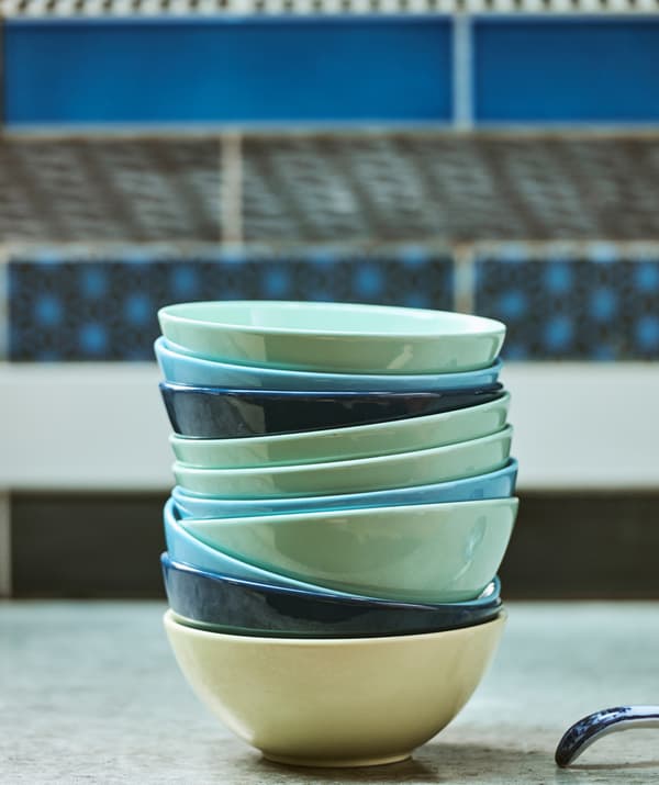 Sur un plan de travail en béton, devant des carrelages muraux bleus à motifs, une pile de bols dans des tons turquoise et bleus clairs et foncés.