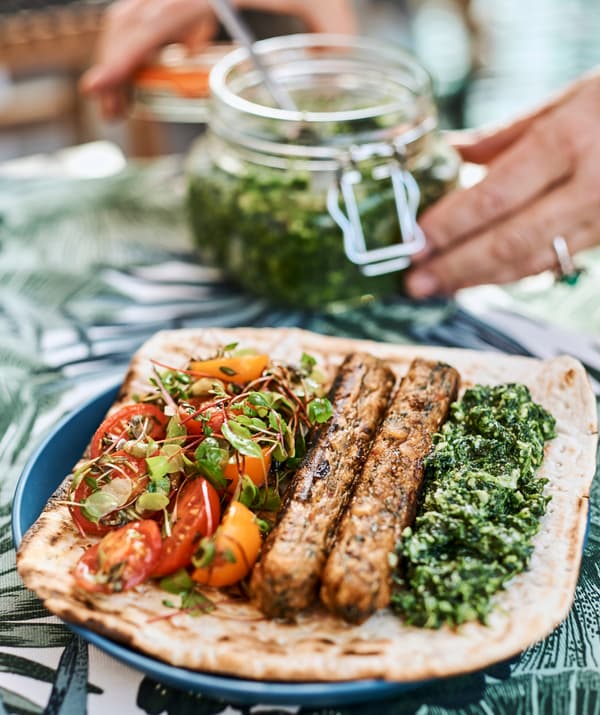 Sur une assiette bleue, un pain plat avec du pesto, des saucisses et de la salade de tomates; on voit des mains qui referment un bocal de pesto.