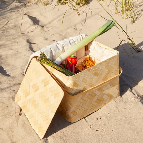 Sur une plage de sable, un panier fraîcheur KÅSEBERGA en bambou tissé et revêtement intérieur en coton contenant différents légumes.
