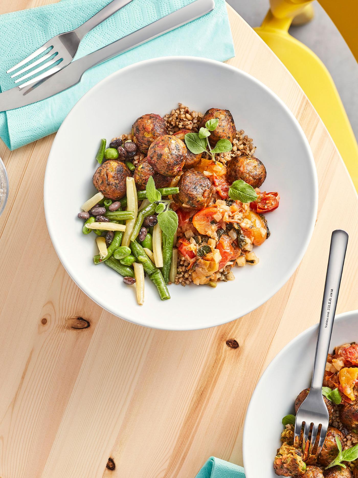 Sur une table en bois, des serviettes de table, des couverts et deux assiettes blanches garnies de boulettes végétales et de légumes cuits multicolores.