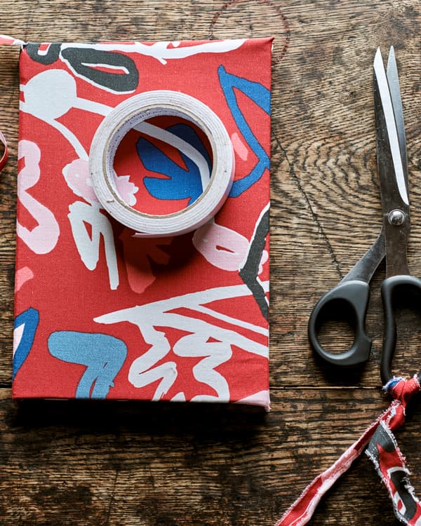 Sur une table en bois rustique, un livre recouvert d’un tissu imprimé rouge avec un rouleau de ruban adhésif posé dessus et une paire de ciseaux à côté.