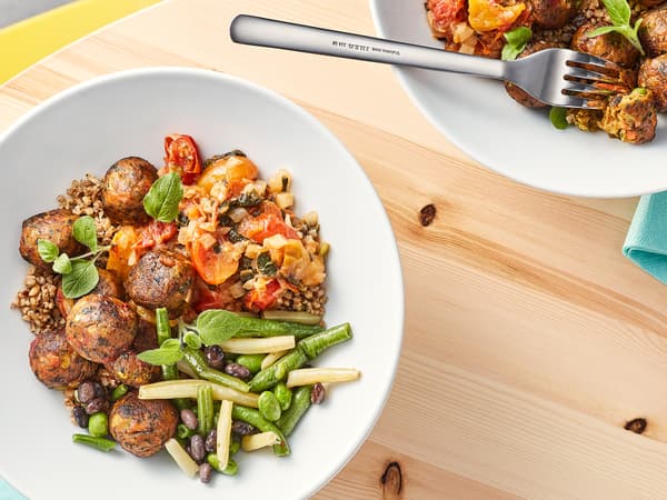 Sur une table en bois sont posées une fourchette et deux assiettes blanches garnies de boulettes végétales, d’herbes aromatiques et de légumes cuits multicolores.
