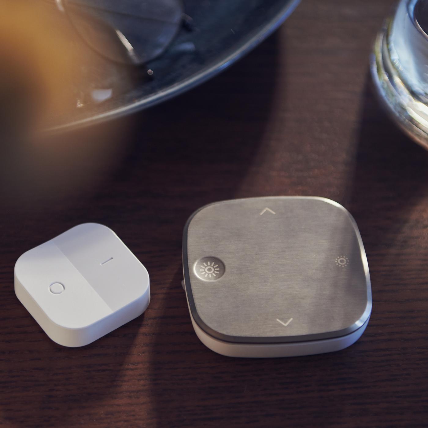 Sur une table en bois teinté foncé, un variateur d’intensité sans fil TRÅDFRI blanc et une télécommande STYRBAR en acier inoxydable.
