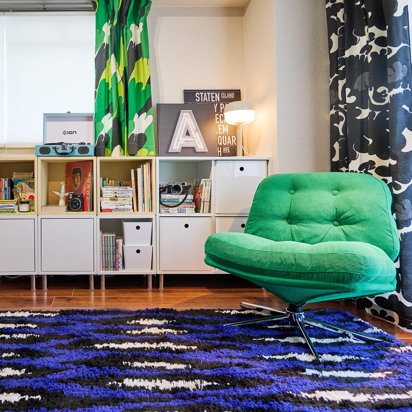 ストライプ柄のラグの上に緑の回転椅子が置かれた、棚と柄入りカーテンのあるカラフルな部屋。