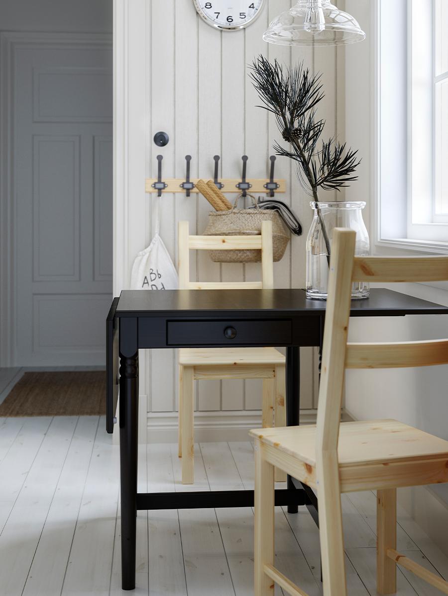 Svart klaffebord med skuff, to furustoler, knaggrekke, veggklokke og taklampe i glass. 