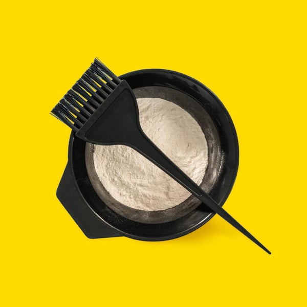 Svart skål med vitt pulver och en svart appliceringspensel, mot gul bakgrund.