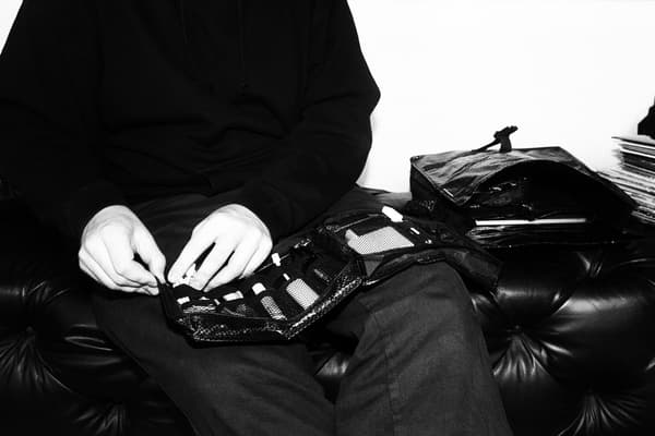 Svartvit bild: en person, som man inte ser huvudet på, sitter på en skinnsoffa och fixar med en mindre svart väska. Bredvid ligger ytterligare en väska