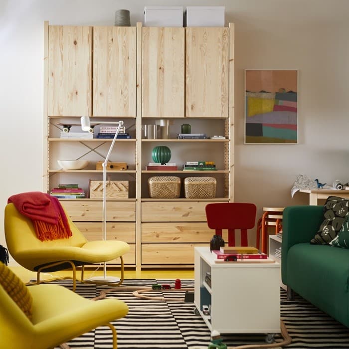 Svetainėje stovi natūralaus medžio daiktų laikymo sistema, žalia sofa, geltonas fotelis su raudonu užtiesalu, baltas kavos staliukas, patiestas dryžuotas kilimas, o ant sienos kabo spalvingas paveikslas. Baldai ir detalės kuria šiltą, jaukią atmosferą.