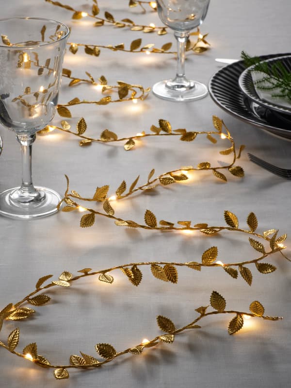 Svetelná reťaz STRÅLA so zlatými lístkami je na bielom obruse medzi pohármi na víno.