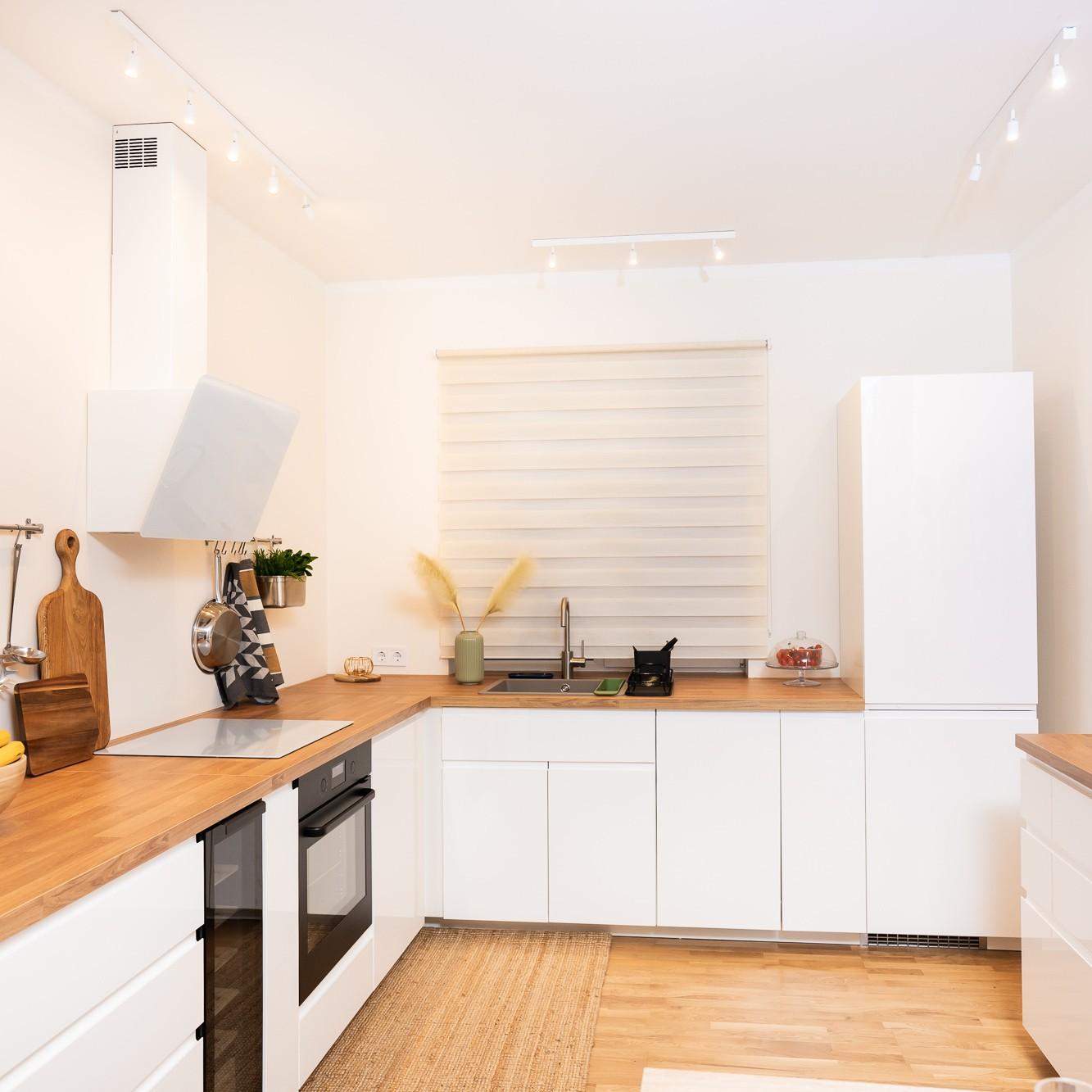 Светлая современная кухня Г-образной формы с белыми фасадами и деревянными столешницами; на столешнице — кухонные принадлежности и растения, создающие аккуратный и просторный вид.