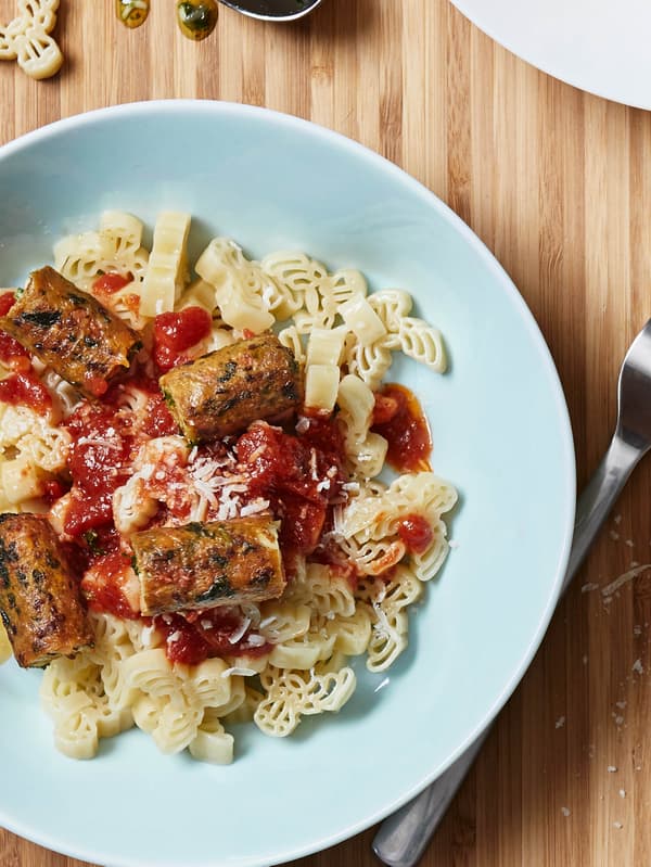 Světle tyrkysový talíř s porcí těstovin BÄSTISAR a opečenými zeleninovými hot dogy KORVMOJ s kečupem a sýrem.