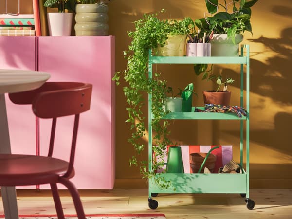 Světle zelený vozík NISSAFORS plný rostlin v květináči stojí před žlutou stěnou.