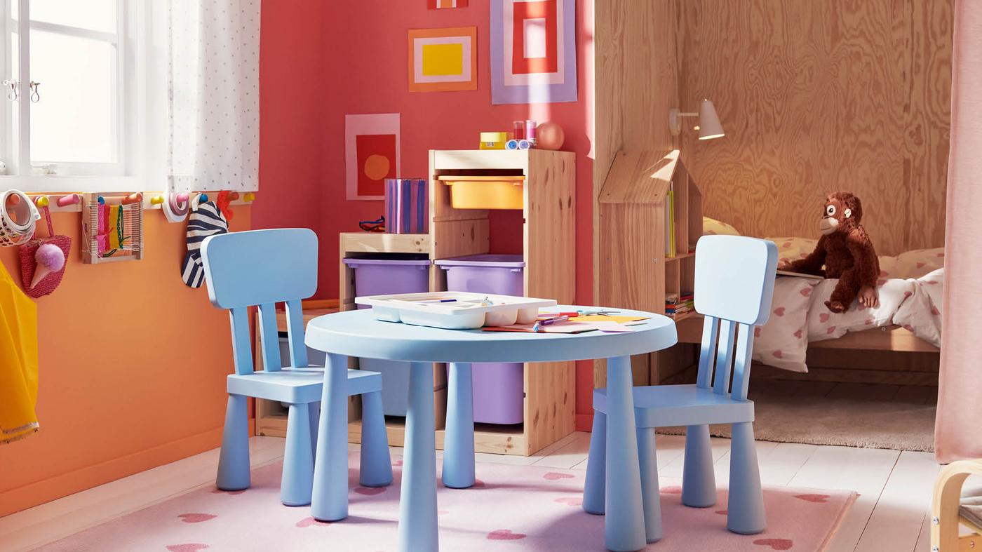 Svetlomodrý detský stôl a dve stoličky MAMMUT, za nimi stojí úložný diel TROFAST s fialovými, modrými a žltými škatuľami.