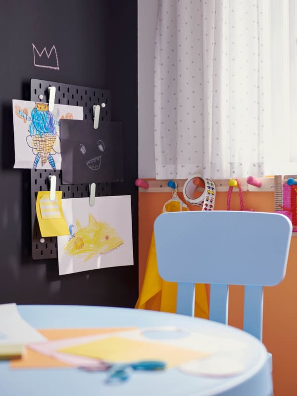 Svetlomodrý detský stôl a stolička MAMMUT, za nimi na stene visí čierny dierkovaný panel SKÅDIS, na ktorom sú bielymi štipcami prichytené obrázky.
