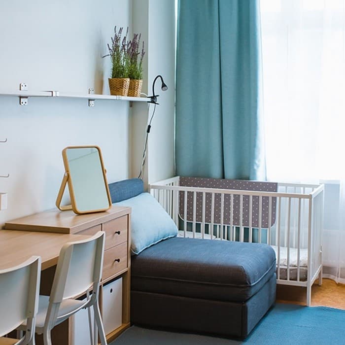 Šviesus, modernaus stiliaus miegamasis su mėlynu išskleidžiamuoju foteliu šalia baltos kūdikio lovelės, staliuku su veidrodžiu ir kėde bei šviesiai mėlynomis užuolaidomis.