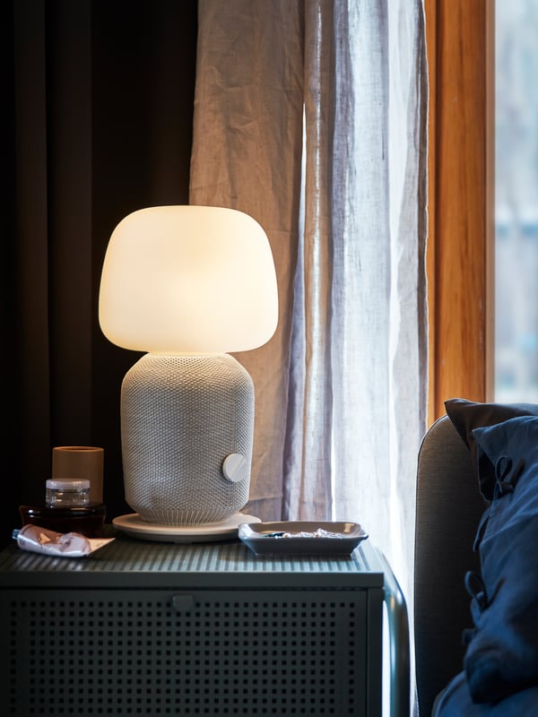 Svietiaca biela lampa SYMFONISK s WiFi reproduktorom umiestnená na zelenom stolíku/skrinke vedľa postele.