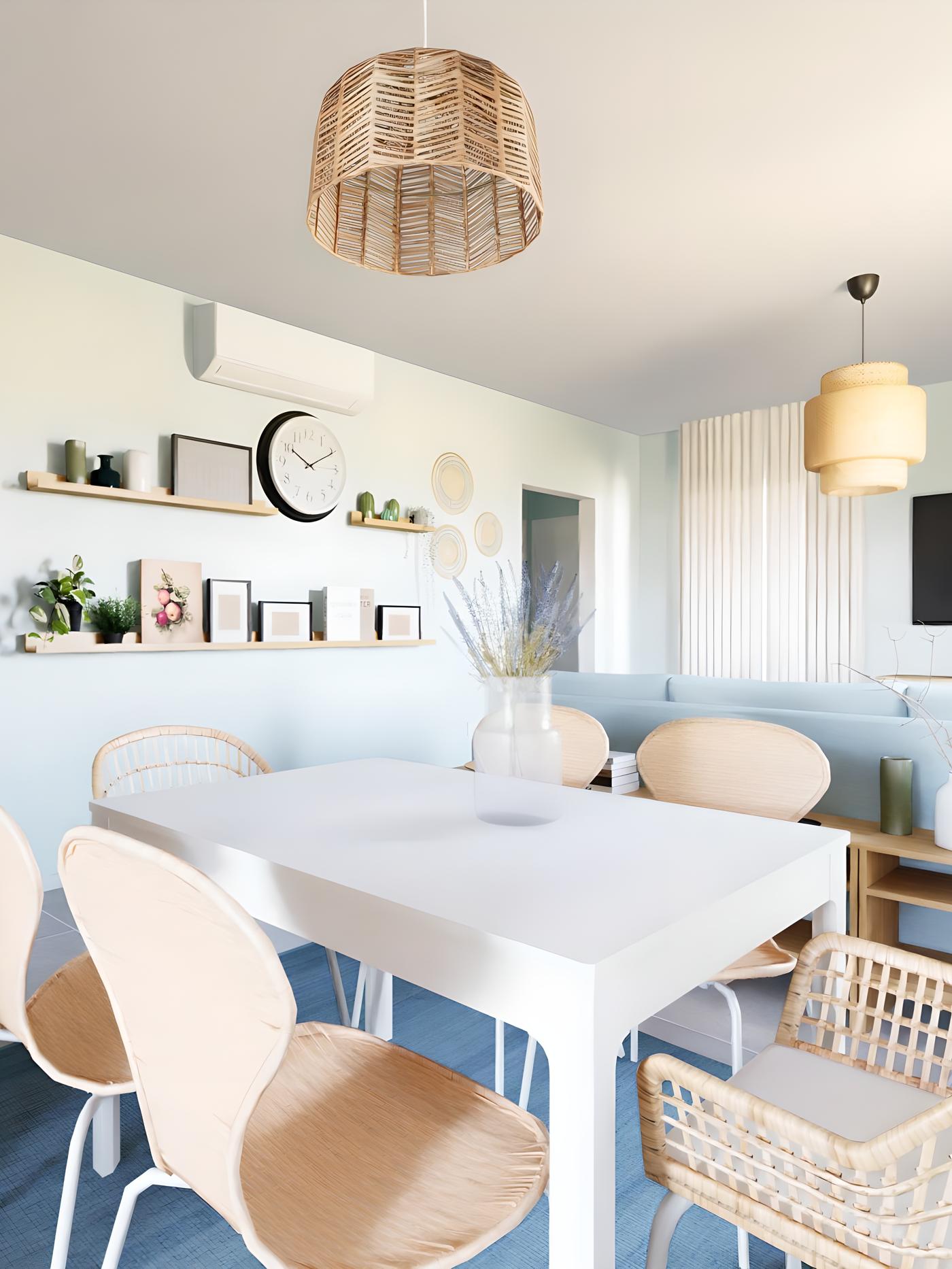 Svijetla blagovaonica s bijelim stolom, stolicama od ratana, visećim lampama, policom s biljkama, knjigama i satom; na stolu vaza s cvijećem, u pozadini plavi kauč i TV na zidu.