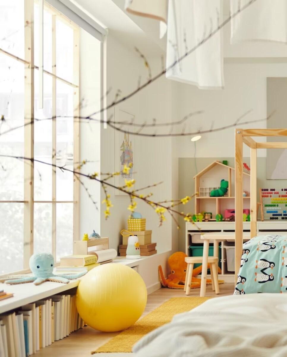 Svijetla dječja soba s drvenim krevetom, stolom, policama za knjige i žutom loptom. Na prozoru su grane s cvjetovima.