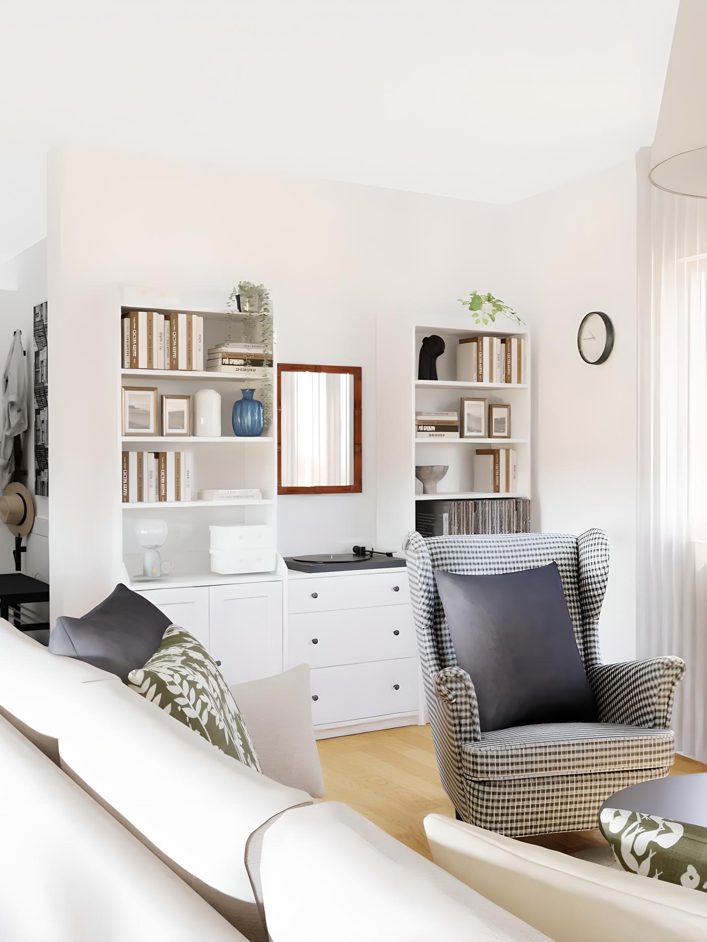 Svijetla dnevna soba s bijelim zidovima, policama za knjige, naslonjačem s crnim jastukom, ogledalom, satom na zidu te knjigama, biljkama i ukrasima na policama.