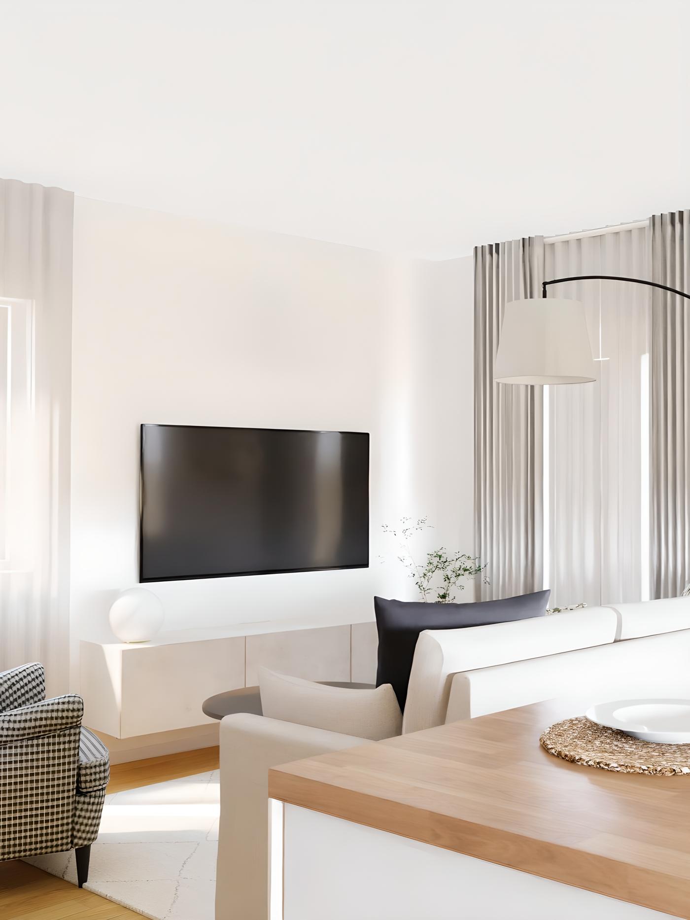 Svijetla dnevna soba s bijelim zidovima, sivom sofom, naslonjačem s uzorkom, drvenim stolom, ravnim televizorom na zidu i velikim prozorima sa svijetlim zavjesama.