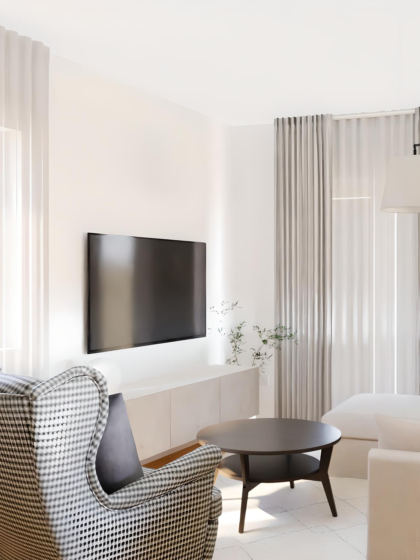 Svijetla dnevna soba s crnim TV-om na zidu, bijelim ormarićem, drvenim stolićem, foteljom s uzorkom i bijelom sofom, osvijetljena prirodnim svjetlom kroz prozirne zavjese.