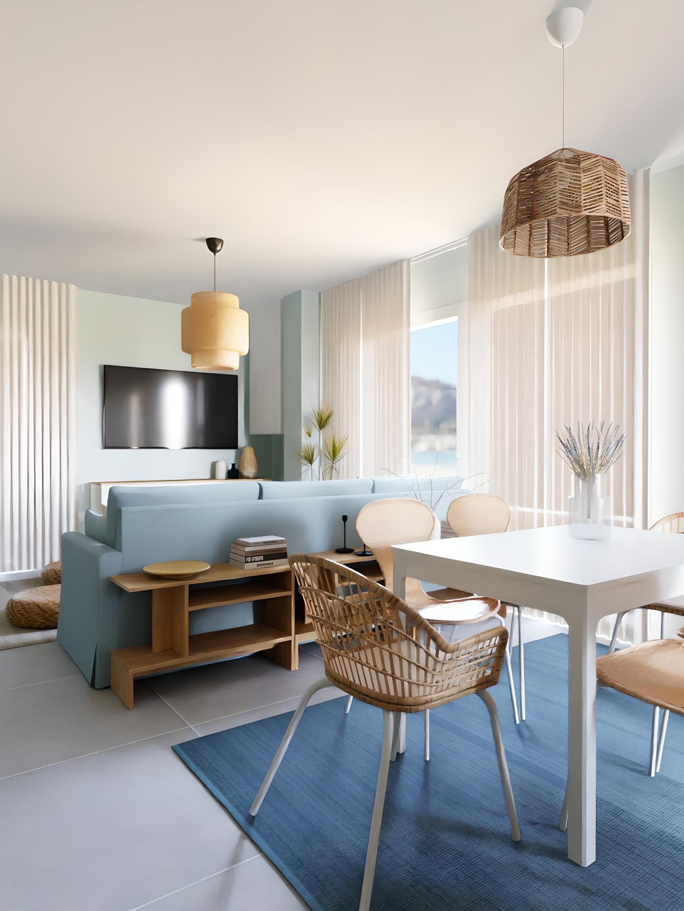 Svijetla dnevna soba s plavim kaučem, bijelim stolom, stolicama od ratana, plavom prostirkom, visećim lampama, velikim prozorima, televizorom na zidu i vazom s cvijećem na stolu.
