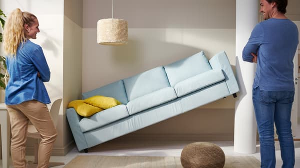 Svijetloplava sofa nagnuta je pod kutom i naslonjena na zid između dvije osobe koje je promatraju; na sofi se nalazi žuti jastuk, a prostor je uređen svijetlim tepihom, pletenom stolicom i visećom lampom.